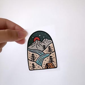 May include: A sticker featuring a mountain landscape with a red sun, stars, a river, trees, and a tent. The sticker is in an arch shape.