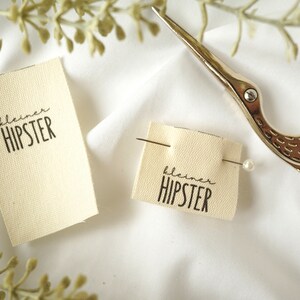 Può includere: Due etichette in tessuto bianco con il testo "kleiner HIPSTER" stampato in nero. Nell'immagine sono presenti anche un paio di forbici argentate con un design a forma di uccello e uno spillo con una testa di perla bianca.
