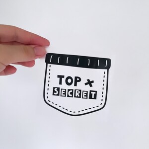 Peut inclure: Autocollant noir et blanc d'une poche avec les mots "TOP SECRET" à l'intérieur. La poche a un design cousu.
