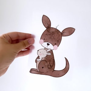 Può includere: Un'illustrazione ad acquerello di un canguro marrone che tiene un piccolo canguro joey marrone nella sua tasca. Il canguro ha le guance rosa e un piccolo cuore sul petto.