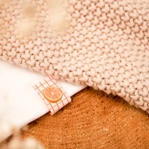 Peut inclure: Un t-shirt blanc avec un petit graphique de fruit d'agrumes orange sur une étiquette en tissu à carreaux rose et blanc. Le t-shirt est posé sur une surface en bois brun avec un tissu tricoté beige sur le dessus.