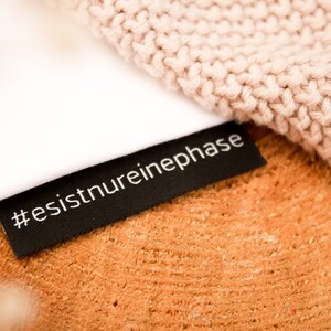Peut inclure: Une étiquette en tissu noir avec du texte blanc qui dit "#esistnureinephase" sur une surface en bois brun clair. Un tissu tricoté rose clair est partiellement visible en arrière-plan.