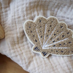 Könnte beinhalten: Ein gestickter Muschelflicken in Beige und Braun mit weißem Umriss. Der Patch befindet sich auf einem beigefarbenen Stoffhintergrund.