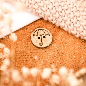 Könnte beinhalten: Ein Holzknopf mit einem Schirm-Design, der auf einem Holzstück liegt. Der Knopf ist braun mit einer schwarzen Schirm-Illustration. Ein gestrickter beiger Stoff und weißes Tuch befinden sich im Hintergrund. Getrocknete Blumen sind im Vordergrund.