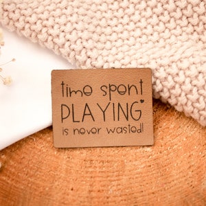 May include: A brown leather tag with the text "time spent PLAYING is never wasted!" on a white and beige knitted fabric background.