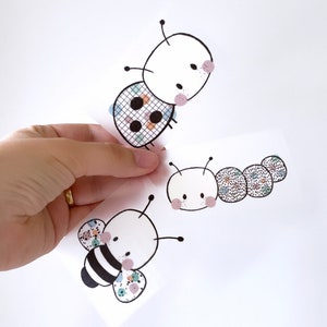 May include: Three colourful stickers featuring a bee, a ladybug, and a caterpillar. Each sticker has a black outline and is decorated with small, colourful dots.