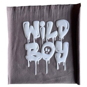 Op de afbeelding: Een bruine stoffen tas met een witte afbeelding van de woorden "WILD BOY" in een druppelende lettertype stijl. Een schedel afbeelding is gecentreerd in het woord "BOY".