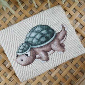 Op de afbeelding: Een bruine en groene schildpad met een schild op een witte stoffen achtergrond. De schildpad is afgebeeld in een cartoon-stijl met een glimlachend gezicht.