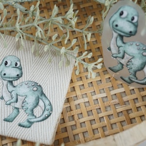 Könnte beinhalten: Zwei grüne Dinosaurier-Illustrationen, eine auf einem durchsichtigen Aufkleber und eine auf einem beigefarbenen Stoff-Patch. Beide Dinosaurier haben weiße Punkte und große Augen.