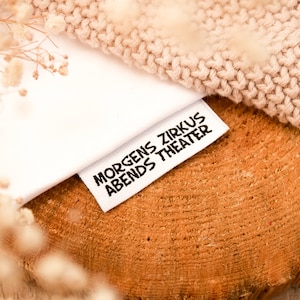 May include: White fabric label with black text that reads "Morgens Zirkus Abends Theater". The label is on a wooden surface with a beige knitted fabric in the background.