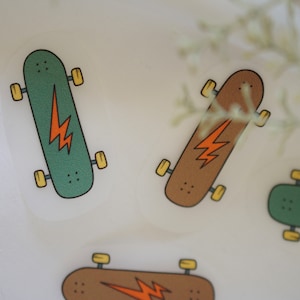 Peut inclure: Trois illustrations de planches à roulettes avec des motifs d'éclairs. Les planches à roulettes sont en vert, marron et marron avec des roues jaunes.