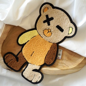 Könnte beinhalten: Ein Plüsch-Teddybär-Aufnäher mit schwarzem Umriss, mit einem Kreuz für ein Auge und einem geschlossenen Auge. Der Bär hat einen beigefarbenen Kopf, ein gelbes Ohr, einen braunen Körper und eine hellbraune Brust. Der Aufnäher liegt auf einem Holztablett.