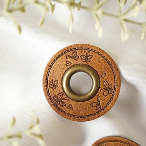 Può includere: Un bottone in pelle marrone con un occhiello in ottone e un design decorativo di farfalle e cuori.