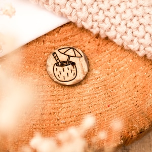 Könnte beinhalten: Ein Holz-Knopf mit einem braunen Kokosnuss-Design und einem schwarzen Regenschirm und Strohhalm. Der Knopf liegt auf einer Holzoberfläche.