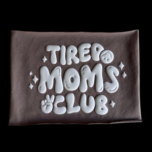 May include: A brown t-shirt with white text that reads "TIRED MOMS CLUB" in a playful, bubbly font. The text is raised and has a 3D effect.