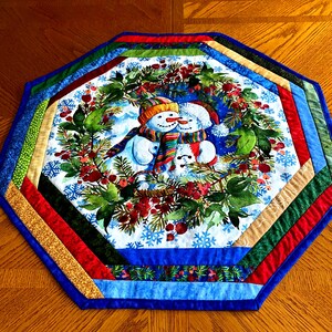 May include: An octagonal Christmas table runner featuring a snowman design. The center shows two snowmen in a wreath of greenery and red berries. The border has a patchwork design with blue, green, red, and gold fabrics. The runner is placed on a wooden table.