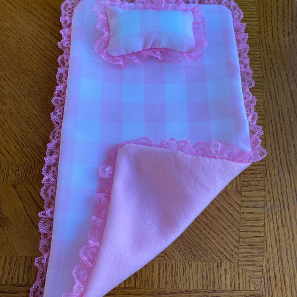 May include: A pink and white gingham doll bed set with a matching pillow and blanket. The set is trimmed with pink lace. The blanket is folded over the bed, revealing a soft pink fleece lining. The set is designed for a doll.