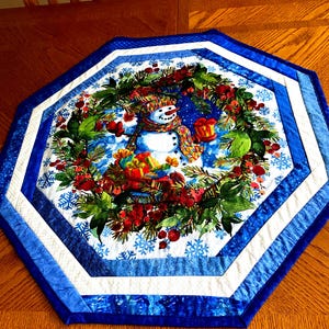May include: An octagonal Christmas table runner featuring a snowman in a wreath of holly and berries. The runner has a blue and white border with snowflake accents, creating a festive winter scene.