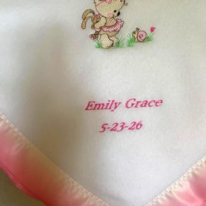 May include: A white baby blanket with a pink satin border featuring an embroidered design of a teddy bear holding a small rabbit, a snail, and the text Emily Grace 5-23-26 in pink thread. The blanket has a soft texture and a triangular corner.