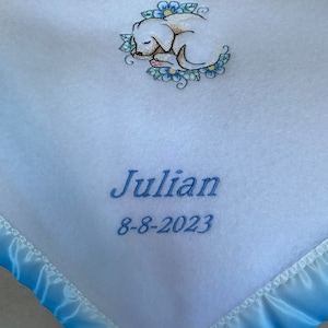 May include: White baby blanket with a light blue satin trim. The blanket features an embroidered sleeping puppy surrounded by blue and green flowers. The name "Julian" and the date "8-8-2023" are embroidered in light blue.