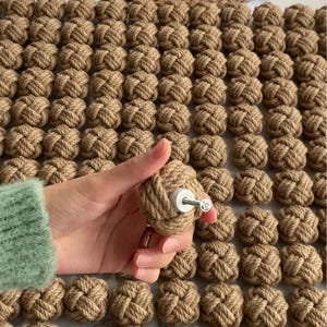 K&ouml;nnte beinhalten: Nahaufnahme von dekorativen Seilknoten, von denen einer in der Hand gehalten wird, mit einem Knaufdesign. Die Knoten sind naturfarben braun, und der Knauf hat eine wei&szlig;e Basis und eine silberne Schraube. Ein kleiner Blumentopf und eine gewebte Matte befinden sich im Hintergrund.