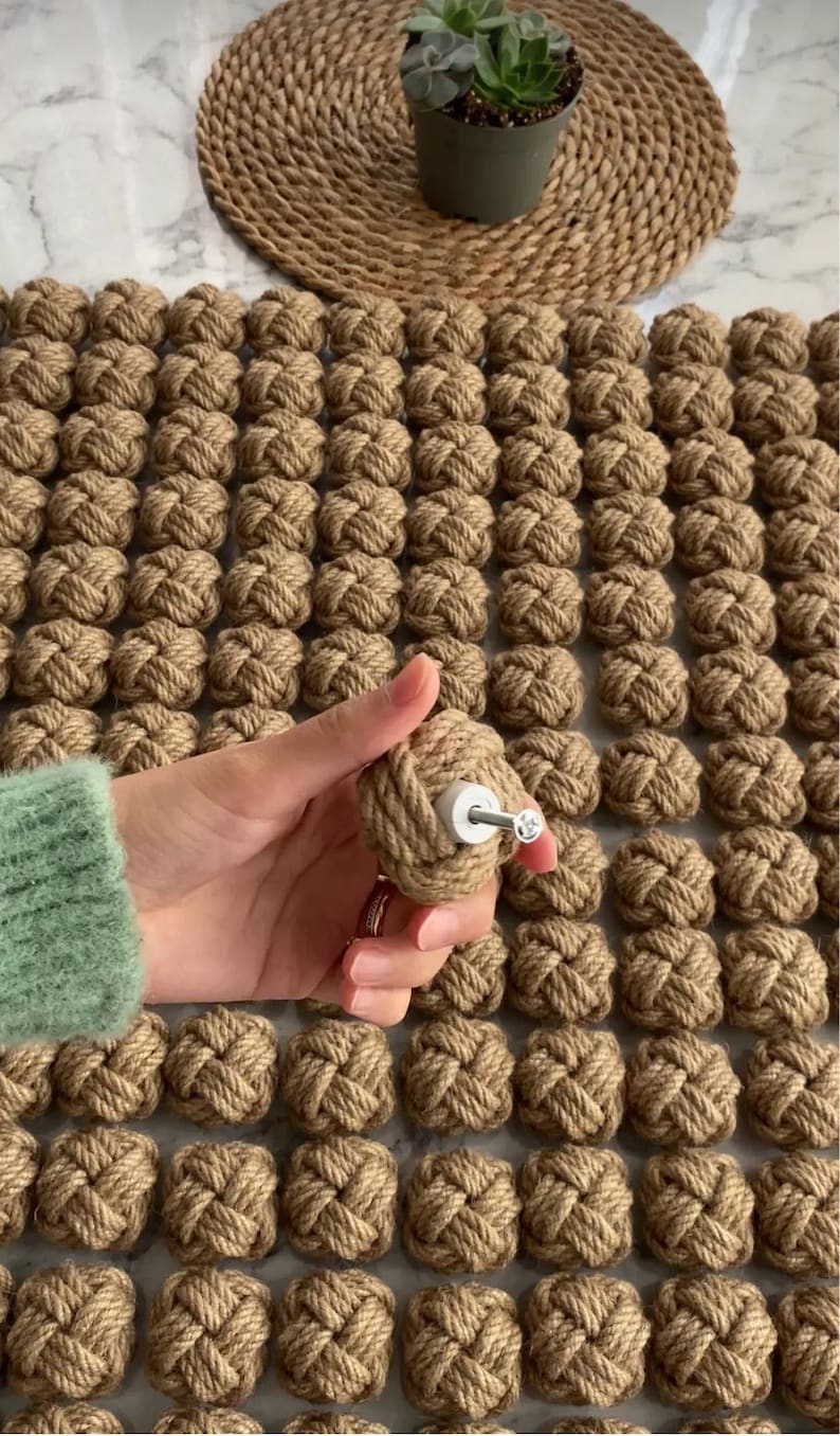 K&ouml;nnte beinhalten: Nahaufnahme von dekorativen Seilknoten, von denen einer in der Hand gehalten wird, mit einem Knaufdesign. Die Knoten sind naturfarben braun, und der Knauf hat eine wei&szlig;e Basis und eine silberne Schraube. Ein kleiner Blumentopf und eine gewebte Matte befinden sich im Hintergrund.