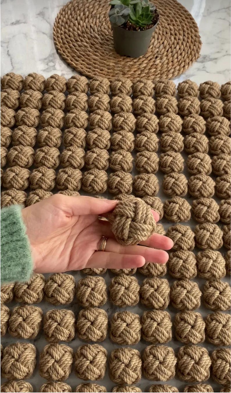 K&ouml;nnte beinhalten: Eine Anordnung von handgebundenen, braunen Seilknoten in einem Rastermuster. Ein Knoten wird in der Hand gehalten, um das aufwendige Webdesign zu zeigen. Im Hintergrund befindet sich eine kleine Topfpflanze auf einem gewebten Platzset.