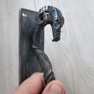 May include: Hand-forged metal hook with a ram's head design. The hook has a rectangular mounting plate with two holes. The hook is twisted and curved, with a detailed ram's head. The metal has a dark, textured finish. Suitable for hanging items.