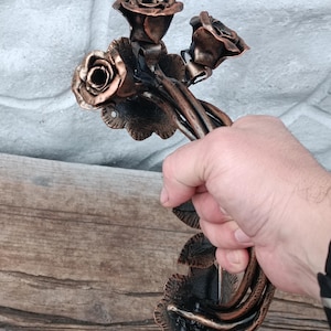Può includere: Una maniglia per porta in metallo color bronzo, realizzata a mano, con un design a rosa. La maniglia presenta tre fiori di rosa e diverse foglie. La maniglia è tenuta da una mano, che mostra gli intricati dettagli della lavorazione del metallo.