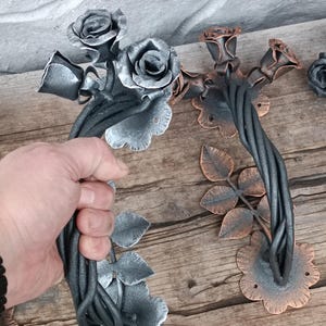 May include: Hand-forged metal door handles with a floral rose design. The handles are dark gray with silver and copper accents. The rose blooms and leaves are intricately detailed, adding a touch of elegance. The handles are mounted on a wooden surface.