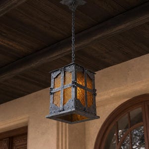 Pode incluir: Uma luminária pendente retangular em ferro forjado com painéis de vidro âmbar. A lanterna é suspensa por uma corrente e um suporte de teto. O design apresenta metalurgia rebitada e um topo decorativo, adequado para uso externo ou interno.