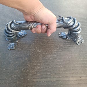 Peut inclure: Une poignée de porte décorative en métal argenté avec un design organique. La poignée présente des détails tourbillonnants en forme de vigne et des accents de feuilles. La poignée est tenue dans une main.
