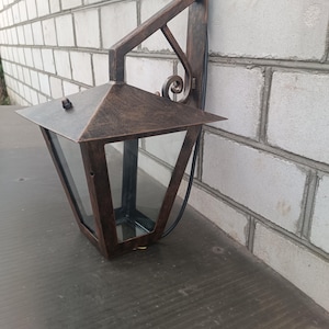 May include: A bronze-coloured outdoor wall lantern with a geometric design. The lantern has a peaked roof and clear glass panels, and is mounted on a decorative bracket. The fixture is designed to be mounted on a brick wall.