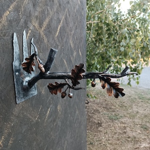 Puede incluir: Un gancho de pared de metal con forma de rama de árbol con hojas y bellotas. El gancho está pintado de negro con hojas y bellotas de color cobre.
