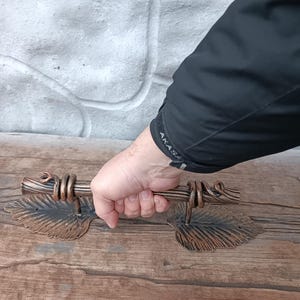 Puede incluir: Manija de puerta de metal color bronce con detalles de hojas y vides. La manija es sostenida por una mano, mostrando el intrincado diseño. La manija tiene un aspecto rústico y antiguo.