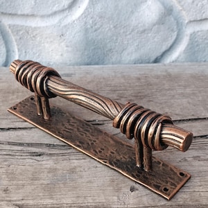 Può includere: Una maniglia per porta color bronzo con base rettangolare. L'impugnatura presenta un design attorcigliato e un'impugnatura cilindrica. Il metallo ha un aspetto rustico e invecchiato, che suggerisce uno stile artigianale o vintage. La maniglia è progettata per una facile installazione.