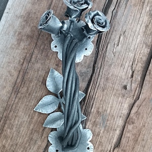 May include: A handcrafted metal door pull featuring a bouquet of roses. The roses, leaves, and stems are intricately detailed in a dark gray finish. The door pull is mounted on a decorative, scalloped base, adding a touch of elegance to any door.