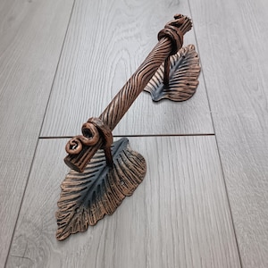 May include: A decorative bronze-colored door handle with a leaf design. The handle has a textured, wood-like appearance and is attached to two stylized leaf-shaped mounts. The metal has a dark bronze finish with hints of black.