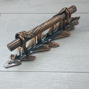 May include: A bronze-coloured towel bar with a twisted handle and leaf-shaped supports. The metal bar has a rustic design, suitable for a bathroom or kitchen. The dimensions are approximately 25 cm in length.