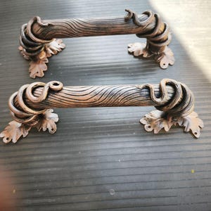 Puede incluir: Dos tiradores de armario en tono bronce. Cada tirador tiene un centro texturizado con aspecto de madera con detalles arremolinados en forma de vid en cada extremo. Los tiradores están montados sobre bases en forma de hoja.