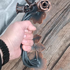 Peut inclure: Une sculpture en métal artisanale de roses et de feuilles en bronze et noir. La conception florale comprend trois roses et plusieurs feuilles, avec une tige torsadée. La sculpture est tenue dans une main.
