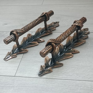 Puede incluir: Dos manijas de puerta de color bronce. Cada manija presenta un diseño de barra retorcida con un detalle de nudo, montada sobre una base en forma de hoja. Las manijas son de metal y tienen una apariencia rústica y antigua.