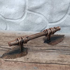 May include: Bronze-colored metal door handle with a vine-like design. The handle features a central bar with coiled accents and leaf-shaped base plates. The handle is approximately 10 inches long.