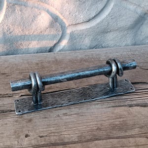 May include: A handcrafted, rustic metal door pull. The pull features a horizontal bar held by two vertical supports with decorative rings. The base is a rectangular plate with hammered texture and mounting holes. The metal has a dark, aged finish.