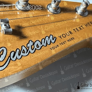 May include: A light brown wooden guitar headstock featuring a silver glitter decal with the word Custom in a cursive script, accompanied by the text YOUR TEXT HERE in a black sans-serif font, with metal tuning pegs and a string tree visible.
