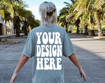Op de afbeelding: Een persoon draagt een lichtblauw T-shirt met de tekst "YOUR DESIGN HERE" in wit. De persoon staat op een geplaveid oppervlak met palmbomen op de achtergrond.