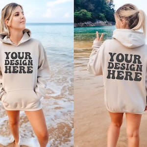 May include: A beige hoodie with the text "YOUR DESIGN HERE" printed on the front and back. The hoodie is being worn by a person standing on a beach.