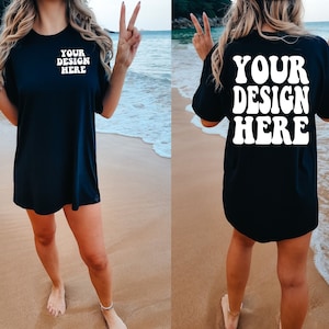 May include: A black t-shirt with the text "YOUR DESIGN HERE" in white on the front and back. The shirt is being worn by a person on a beach.