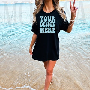 Puede incluir: Una mujer lleva una camiseta negra de gran tamaño con el texto "YOUR DESIGN HERE" en azul claro en la parte delantera. Está de pie en una playa de arena con el océano de fondo.