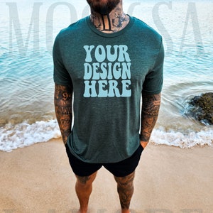 May include: A dark green t-shirt with the text "YOUR DESIGN HERE" in light blue. The shirt is short-sleeved and worn with black shorts. The person is standing on a sandy beach with the ocean in the background.